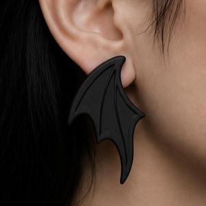 May include: A matte black earring shaped like a stylised bat wing. The earring features defined structural lines that mimic the skeletal structure of a wing, with a pointed tip and curved edges, designed to be worn through an earlobe piercing.