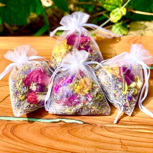 Puede incluir: Cuatro pequeñas bolsas de organza blancas llenas de lavanda seca, pétalos de rosa y otras flores. Las bolsas están atadas con una cinta blanca y están dispuestas sobre una superficie de madera.