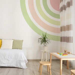 Könnte beinhalten: Ein weißes Bett mit gelben, weißen und grünen Kissen, einer weißen Decke und einer grünen Pflanze in einem weißen Topf. Die Wand hinter dem Bett ist mit einem Regenbogen in Grün-, Rosa- und Weißtönen bemalt. Im Vordergrund steht ein Holztisch mit einer weißen Tischplatte und Holzstühlen.