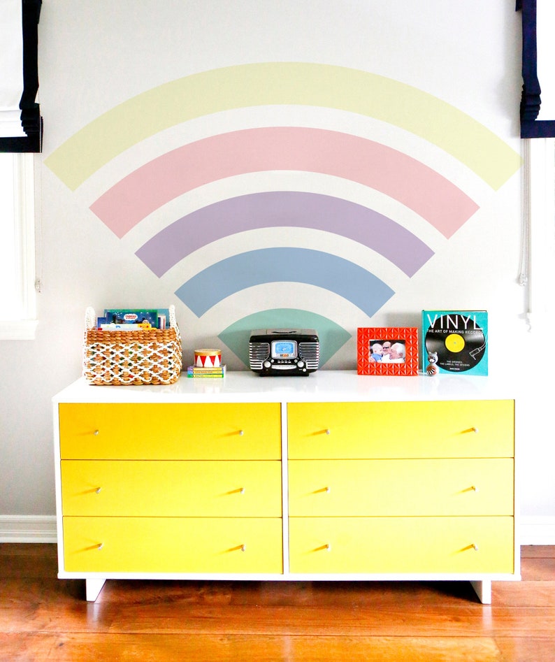 May include: A yellow dresser with six drawers and white trim. The dresser is decorated with a rainbow painted on the wall behind it. The dresser is topped with a vintage radio, a picture frame, a book titled "Vinyl", and a basket of books.