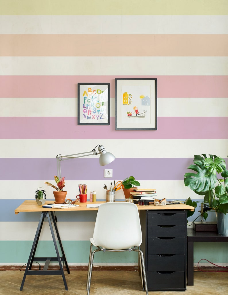 K&ouml;nnte beinhalten: Ein heller Holzschreibtisch mit einem schwarzen Metallgestell und einem wei&szlig;en Stuhl. Der Schreibtisch hat einen schwarzen Schubladenblock und eine Topfpflanze. Die Wand hinter dem Schreibtisch ist mit horizontalen Streifen in Pastellfarben gestrichen. An der Wand h&auml;ngen auch zwei gerahmte Bilder, eines mit einem bunten Alphabet und das andere mit einer einfachen Illustration.