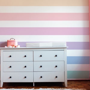 K&ouml;nnte beinhalten: Eine wei&szlig;e Kommode mit sechs Schubladen und schwarzen Kn&ouml;pfen steht vor einer Wand mit horizontalen Streifen in Pastellfarben, darunter Pfirsich, Rosa, Lavendel, Hellblau und Gr&uuml;n.