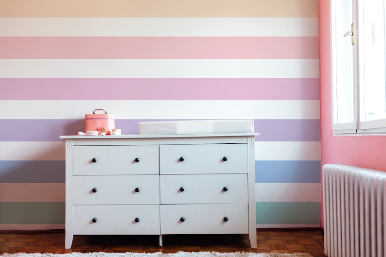 K&ouml;nnte beinhalten: Eine wei&szlig;e Kommode mit sechs Schubladen und schwarzen Kn&ouml;pfen steht vor einer Wand mit horizontalen Streifen in Pastellfarben, darunter Pfirsich, Rosa, Lavendel, Hellblau und Gr&uuml;n.