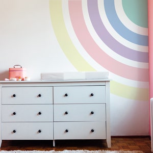 May include: White dresser with six drawers and black knobs. The dresser is in a nursery with a pink and yellow rainbow painted on the wall.