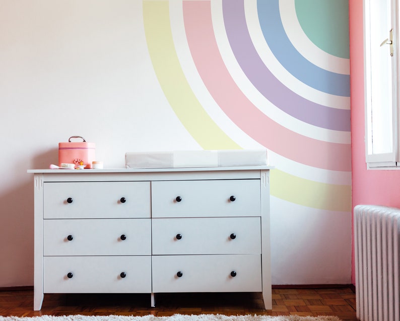 May include: White dresser with six drawers and black knobs. The dresser is in a nursery with a pink and yellow rainbow painted on the wall.