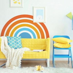May include: A yellow couch with a white throw blanket and a blue patterned pillow sits in front of a wall with a rainbow decal. A blue folding chair with yellow pillows is in the background.