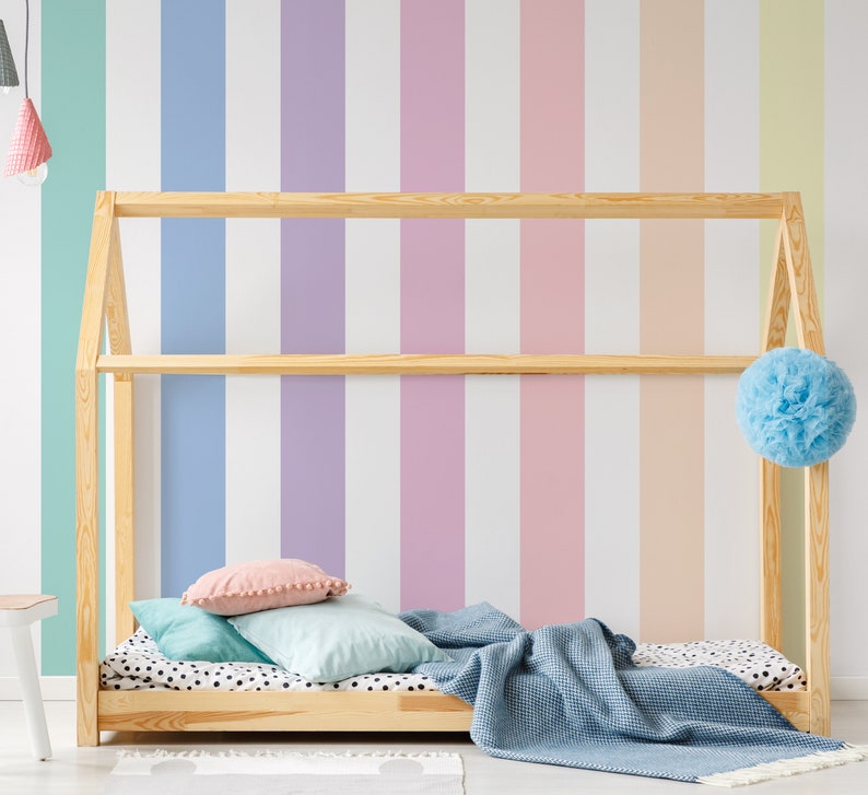 K&ouml;nnte beinhalten: Ein Holzbettgestell mit einer blauen Decke und Kissen vor einer Wand mit pastellfarbenen vertikalen Streifen.
