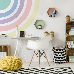 May include: A white desk with a white chair and a yellow knitted ottoman in front of it. The wall behind the desk is painted with a rainbow arc. There are two hexagonal shelves on the wall, one with a clock and the other with a stuffed elephant. A wooden shelf unit with books and other items is on the right side of the image.