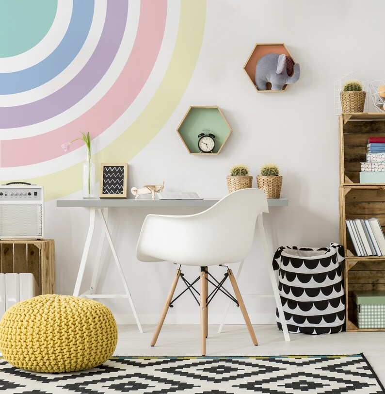 May include: A white desk with a white chair and a yellow knitted ottoman in front of it. The wall behind the desk is painted with a rainbow arc. There are two hexagonal shelves on the wall, one with a clock and the other with a stuffed elephant. A wooden shelf unit with books and other items is on the right side of the image.