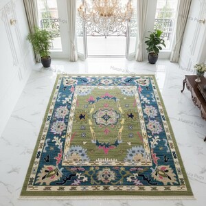 Hand knotted Oushak rug with green center and blue floral border displayed in a bright living room. The wool area rug features traditional Turkish medallion patterns with ivory, pink, and gold accents, placed on a marble floor beneath a chandelier, enhancing a modern luxury interior.