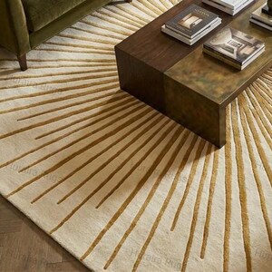 May include: A cream-colored area rug with a sunburst design in shades of gold and brown. The rug is placed in a living room setting, partially under a dark brown coffee table and a green sofa. The rug's pattern radiates outwards from the center.