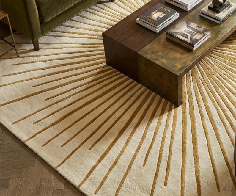 May include: A cream-colored area rug with a sunburst design in shades of gold and brown. The rug is placed in a living room setting, partially under a dark brown coffee table and a green sofa. The rug's pattern radiates outwards from the center.