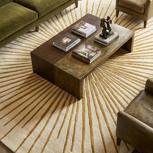 May include: A cream-colored rug with a sunburst design in gold. The rug is in a living room setting with a brown coffee table, green sofa, and leather armchairs. Books and a sculpture are on the table.