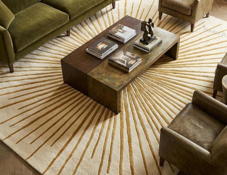May include: A cream-colored rug with a sunburst design in gold. The rug is in a living room setting with a brown coffee table, green sofa, and leather armchairs. Books and a sculpture are on the table.