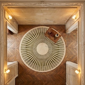 May include: Overhead view of a round rug with a cream-colored center and olive green radiating stripes. The rug is on a wooden floor, with a small wooden table and books visible. The room has ornate architectural details.