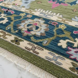 Hand knotted Oushak rug with green center and blue floral border displayed in a bright living room. The wool area rug features traditional Turkish medallion patterns with ivory, pink, and gold accents, placed on a marble floor beneath a chandelier, enhancing a modern luxury interior.