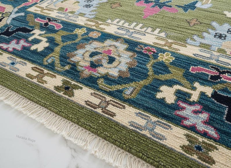 Hand knotted Oushak rug with green center and blue floral border displayed in a bright living room. The wool area rug features traditional Turkish medallion patterns with ivory, pink, and gold accents, placed on a marble floor beneath a chandelier, enhancing a modern luxury interior.