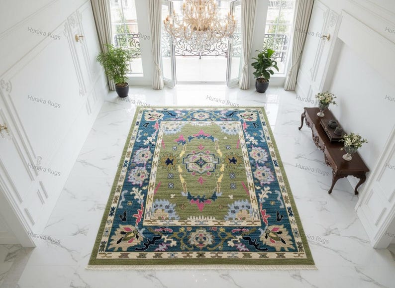 Hand knotted Oushak rug with green center and blue floral border displayed in a bright living room. The wool area rug features traditional Turkish medallion patterns with ivory, pink, and gold accents, placed on a marble floor beneath a chandelier, enhancing a modern luxury interior.