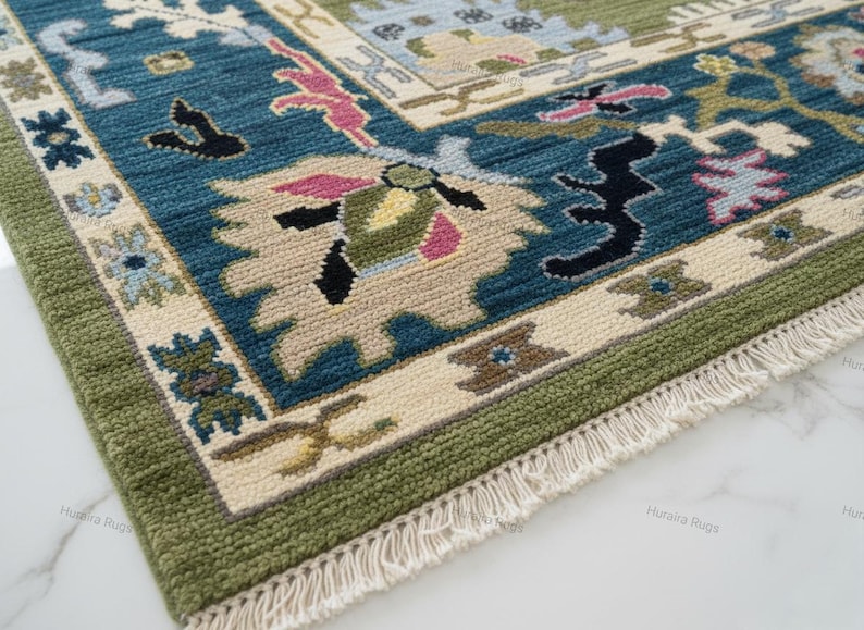 May include: Close-up of a rectangular rug with a detailed pattern. The rug features a teal, beige, and green color scheme with floral and geometric designs. The border is green, and the rug has a fringe.