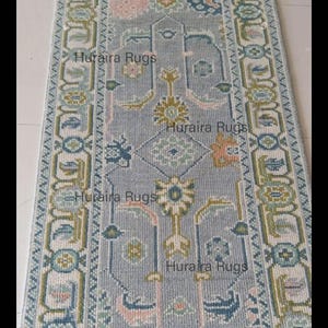 May include: A rectangular, patterned rug with a light blue background and intricate floral and geometric designs in shades of pink, green, and blue. The rug has a border with repeating patterns and the text "Huraira Rugs".