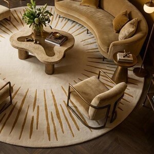 May include: A modern living room with a circular rug featuring a sunburst pattern in beige and gold. The room includes a curved sofa, armchairs, and a unique coffee table. The colour scheme is neutral with warm tones. The rug's diameter is approximately 300 cm.
