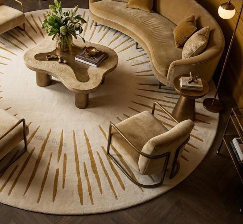 May include: A modern living room with a circular rug featuring a sunburst pattern in beige and gold. The room includes a curved sofa, armchairs, and a unique coffee table. The colour scheme is neutral with warm tones. The rug's diameter is approximately 300 cm.