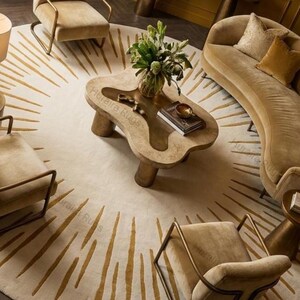 May include: An overhead view of a living room with a large, circular rug with a sunburst pattern in gold and cream. The room features a curved sofa, armchairs, and a distinctive coffee table. The furniture is in beige and gold tones, creating a luxurious and cohesive aesthetic.