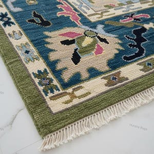 May include: Close-up of a hand-knotted rug with a detailed pattern. The rug features a teal blue field with beige and pink accents, framed by a green border. The design includes floral and geometric motifs, with a fringe along one edge.