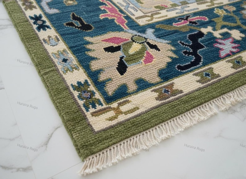 May include: Close-up of a hand-knotted rug with a detailed pattern. The rug features a teal blue field with beige and pink accents, framed by a green border. The design includes floral and geometric motifs, with a fringe along one edge.