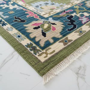 May include: A corner view of a detailed area rug with a traditional design. The rug features a teal, green, and cream color palette with floral and geometric patterns. The edges are finished with a green border and fringe.