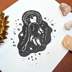 以下が含まれることがあります： 宇宙服を着た犬の白黒の版画で、周りに「The Patron Saint of One Way Trips」という文字が書かれている。