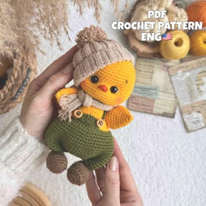 May include: A small, handmade crochet chick toy featuring a bright yellow body, a beige pom-pom beanie, a matching beige scarf, and olive green overalls with two small wooden buttons. The chick has black bead eyes and a tiny orange beak.
