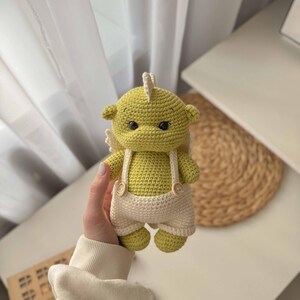 May include: A crocheted green dragon toy wearing white overalls with brown buttons. The dragon has a white crocheted wing on each side.