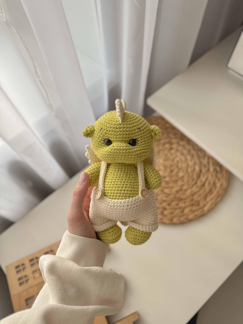 May include: A crocheted green dragon toy wearing white overalls with brown buttons. The dragon has a white crocheted wing on each side.