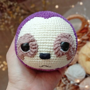 May include: A crocheted sloth toy with purple, white, and brown yarn. The sloth has large eyes and a small nose.