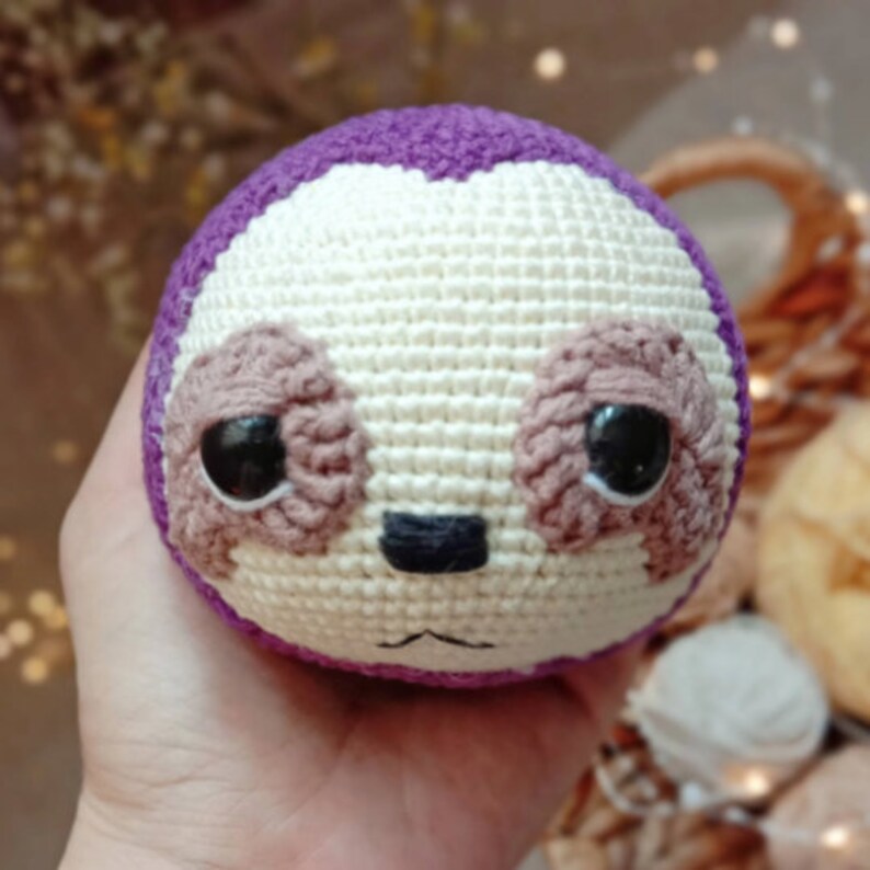 May include: A crocheted sloth toy with purple, white, and brown yarn. The sloth has large eyes and a small nose.