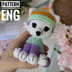 Op de afbeelding: Gehaakte ijsbeerfiguur, wit met zwarte ogen en neus. Het draagt een mintgroene en paarse outfit en hoed. De tekst "PATTERN ENG" staat linksboven. Handgemaakt.