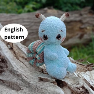Peut inclure: Un jouet amigurumi en forme d'escargot bleu crocheté avec une coquille brune et un motif rayé. L'escargot a un visage mignon avec des yeux noirs et des joues roses.