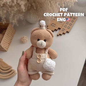 Op de afbeelding: Een gehaakte teddybeer die een witte overall draagt met bruine knopen. De beer heeft een bruine neus en zwarte ogen. De beer houdt een witte pompon op zijn hoofd. PDF CROCHET PATTERN ENG