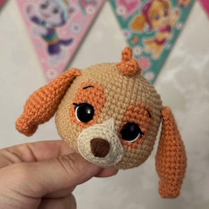 Puede incluir: Cabeza de perro de ganchillo en beige y naranja, con grandes ojos negros y una nariz marr&oacute;n. El perro tiene orejas naranjas y un peque&ntilde;o mech&oacute;n de pelo naranja. El fondo incluye coloridas pancartas triangulares con personajes de dibujos animados.