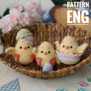 Pode incluir: Três pintinhos amarelos de crochê em uma cesta de vime. Um pintinho está usando um chapéu de crochê em forma de casca de ovo, outro está sentado em uma casca de ovo de crochê, e o terceiro pintinho está segurando um ovo de crochê.