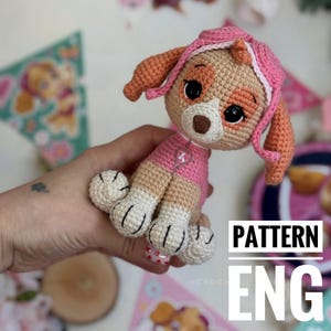 May include: A crocheted Skye puppy toy, in pink and tan. The toy has large black eyes, a pink helmet, and a pink outfit. The text "PATTERN ENG" is visible in the image.