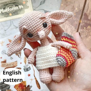 Könnte beinhalten: Häkelpuppe eines Haus-Elfen mit rosa Haut, großen Ohren und einem weißen Kleid. Der Elf hält einen rot-gelb-grauen Häkelschal.