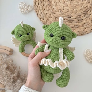 May include: Two crocheted green dragon toys with white wings and a white ruffle. The larger dragon has a wooden ring attached to its arms.