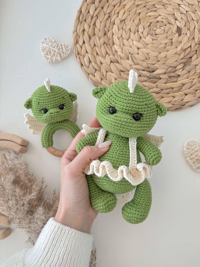 May include: Two crocheted green dragon toys with white wings and a white ruffle. The larger dragon has a wooden ring attached to its arms.