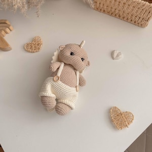 May include: A crocheted stuffed animal with a brown body and white overalls. The animal has a small horn on its head and is lying on a white surface.