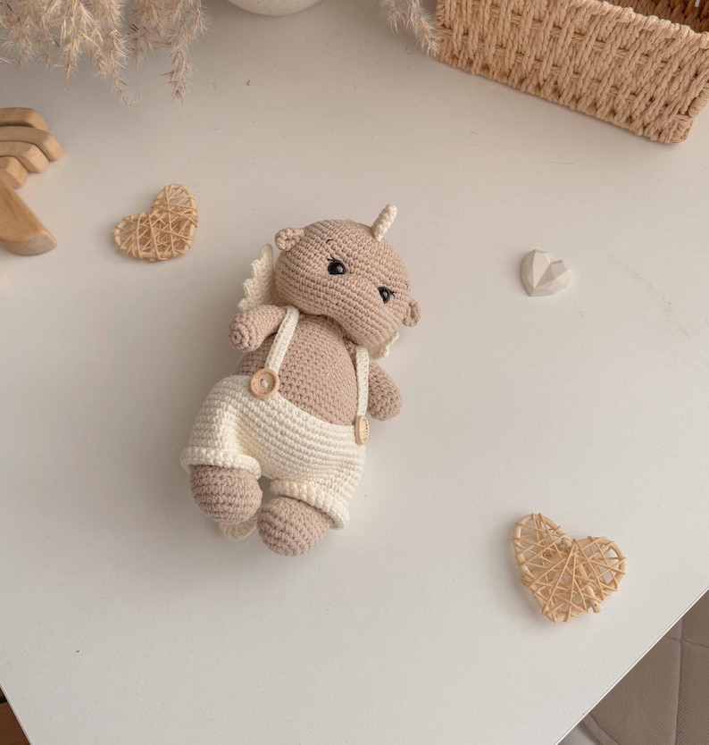 May include: A crocheted stuffed animal with a brown body and white overalls. The animal has a small horn on its head and is lying on a white surface.