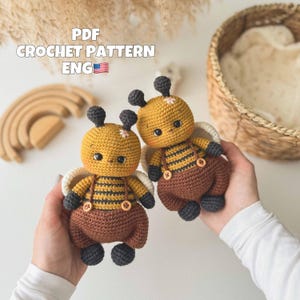 Peut inclure: Deux poupées abeilles crochetées à la main. Les abeilles ont des corps jaunes avec des rayures noires, des salopettes marron et des antennes et pieds noirs. Les poupées ont des ailes blanches. Le texte "PDF CROCHET PATTERN ENG" est visible.