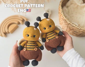 May include: Two handmade crochet bee dolls. The bees have yellow bodies with black stripes, brown overalls, and black antennae and feet. The dolls have white wings and are held in hands. The text "PDF CROCHET PATTERN ENG" is visible.