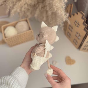 May include: A crocheted plush toy in the shape of a brown and white dragon wearing a white crocheted overall. The dragon has a long tail and a small, pointed snout.